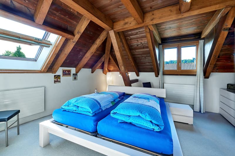 Two beds with blue covers, white bed frame, wooden beams ceiling, radiator, stool, window, small window