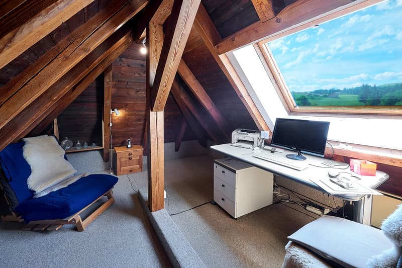 Desk with computer, printer, mouse, books. Blue chair with pillow. Wooden wall, wooden ceiling. Sky view from the window.