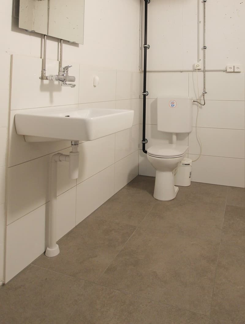 Small white tiled room, white sink, toilet bowl, and small floor-mounted trash bin