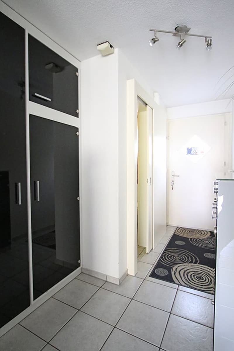 A hallway with a large wardrobe and a door leading to another room. Floor covered with tiles, walls and ceiling in white. A door with a window on it, leading outside.