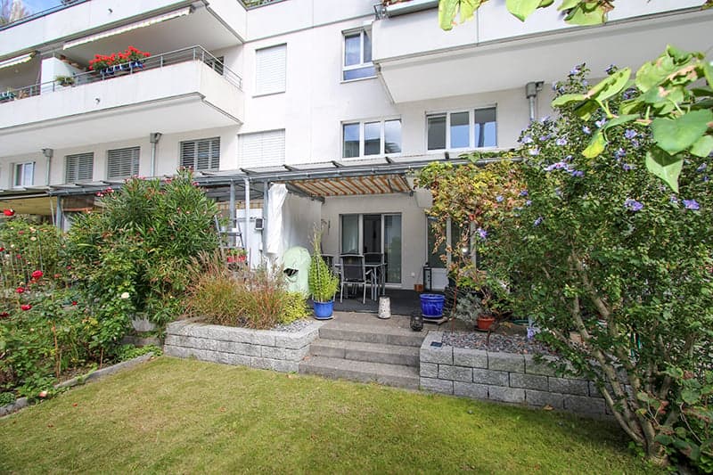A white building with balconies, a garden in the front, a covered seating area with a table and chairs.