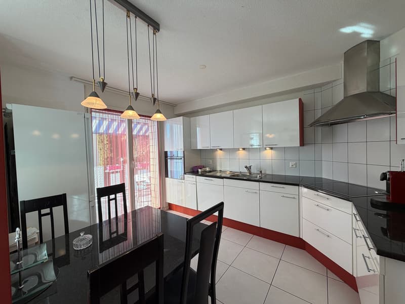 white kitchen with black countertops, white cabinets, island with sink, glass dining table, hanging lamps, white tiles, red glass door