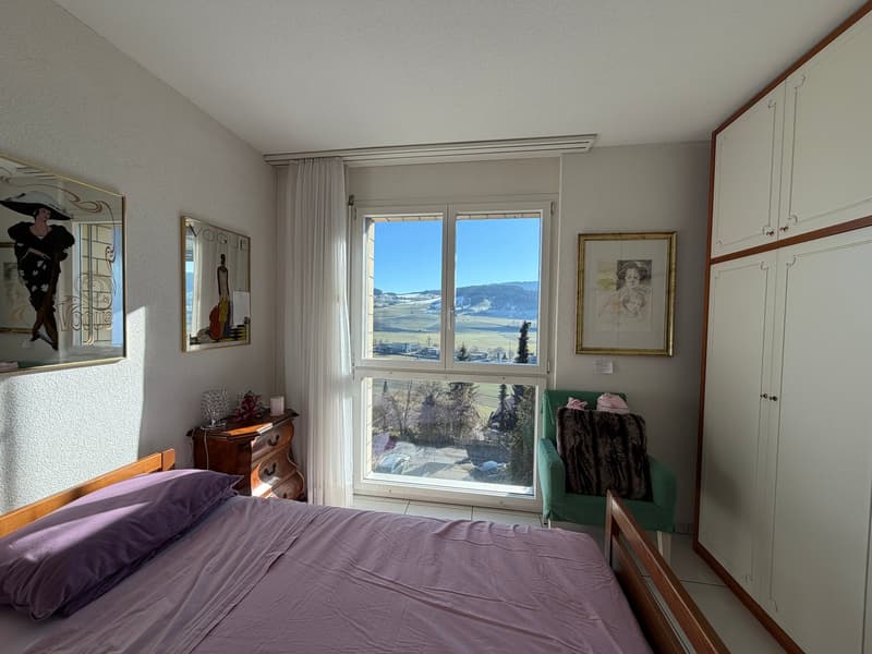 Bedroom with large window, view of mountains, wooden bed, purple pillows, green armchair, wooden cabinet, picture frames, side table