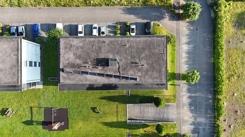 Aerial view of a building with a flat roof, solar panels, and a parking lot with several cars parked.