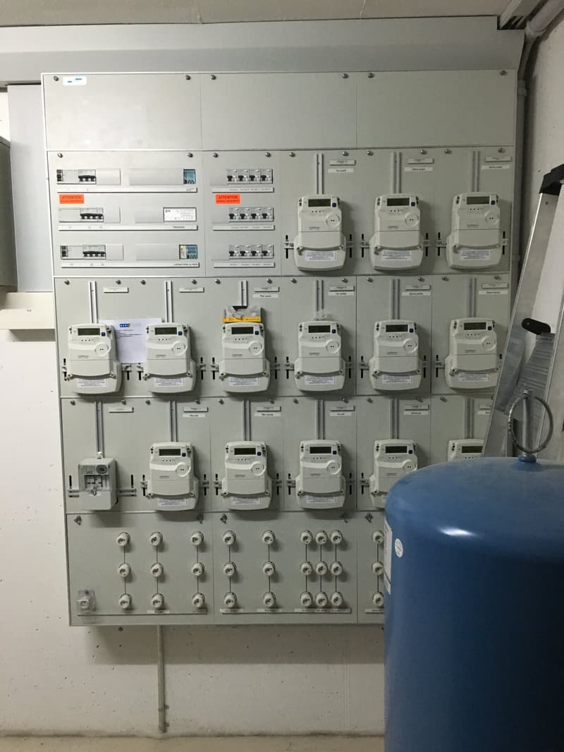 Electrical panel with multiple meters and switches, mounted on a wall, with a blue tank nearby