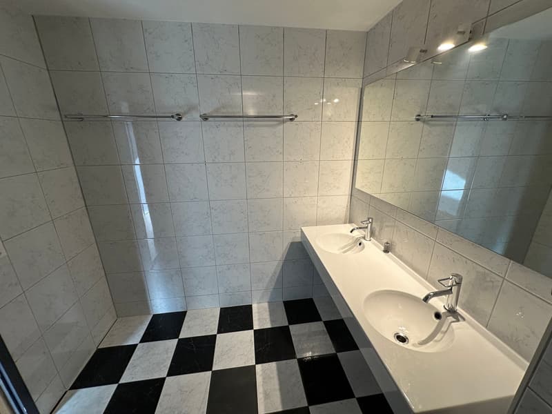 white tile walls, white countertops, black and white checkered floor, two sinks with silver faucets, a large mirror