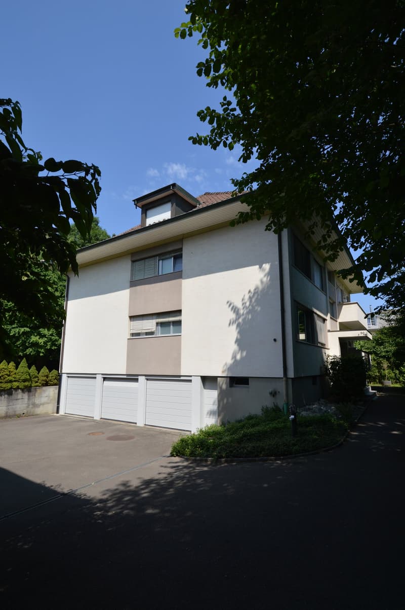 Three story house, white exterior, brown roof, two garages, driveway, shrubs, and trees in the foreground.
