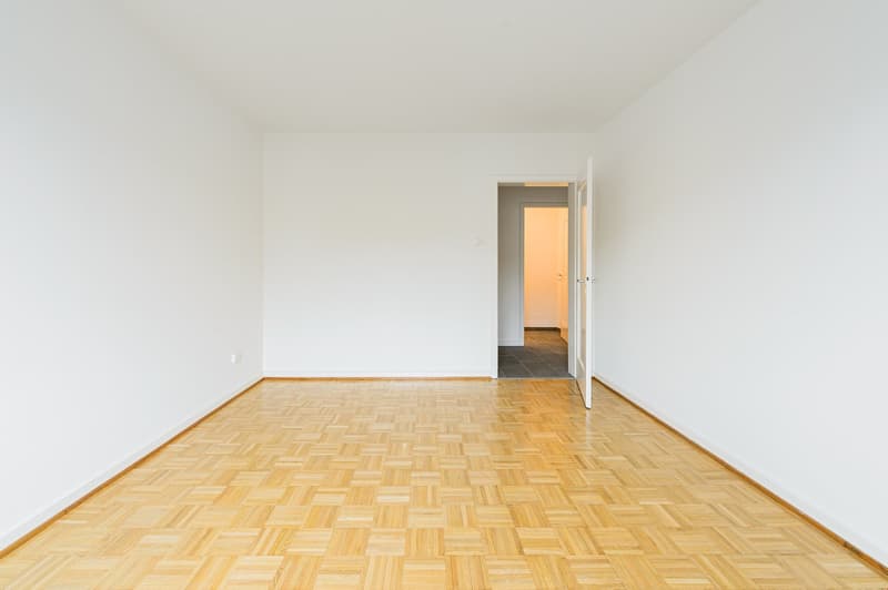 Empty room with wooden parquet floor, white walls, and an open door leading to a smaller room with a tiled floor.