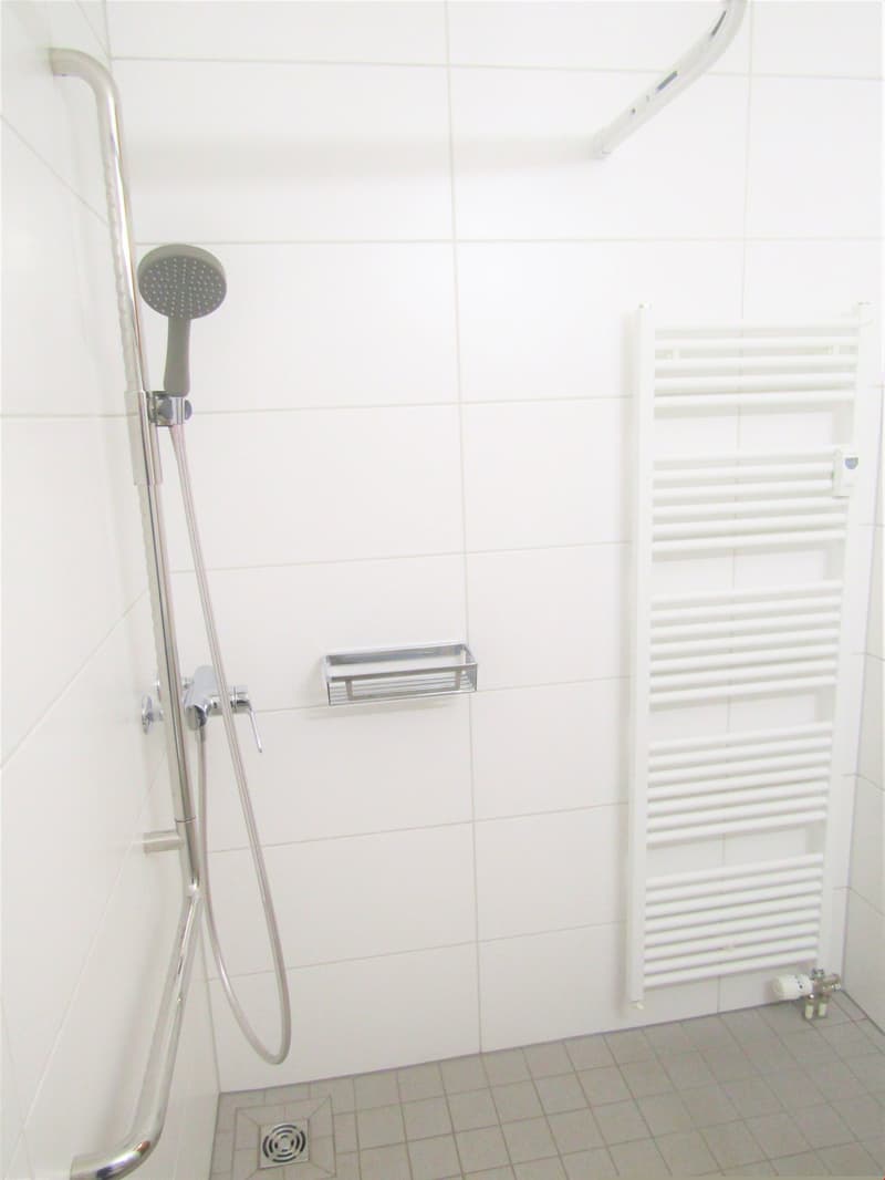 shower area with shower head, faucet, white tiled walls, white radiator, small shelf, shower drainage