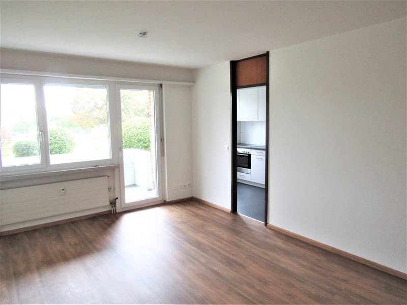 Empty room with sliding glass door leading to balcony, wooden floor, white walls, radiator, electrical outlet, entrance to kitchen