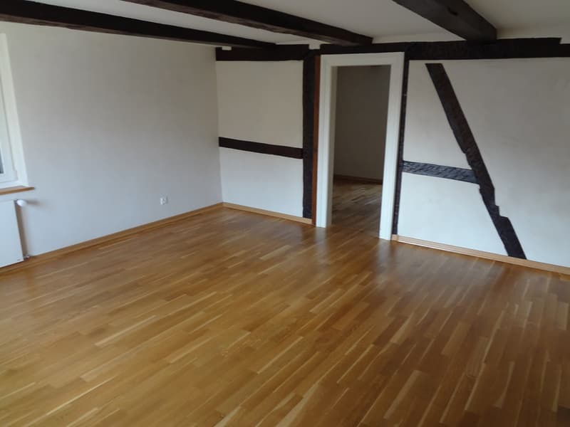 Large room, wooden floor, white walls, exposed wooden ceiling beams, window with radiator