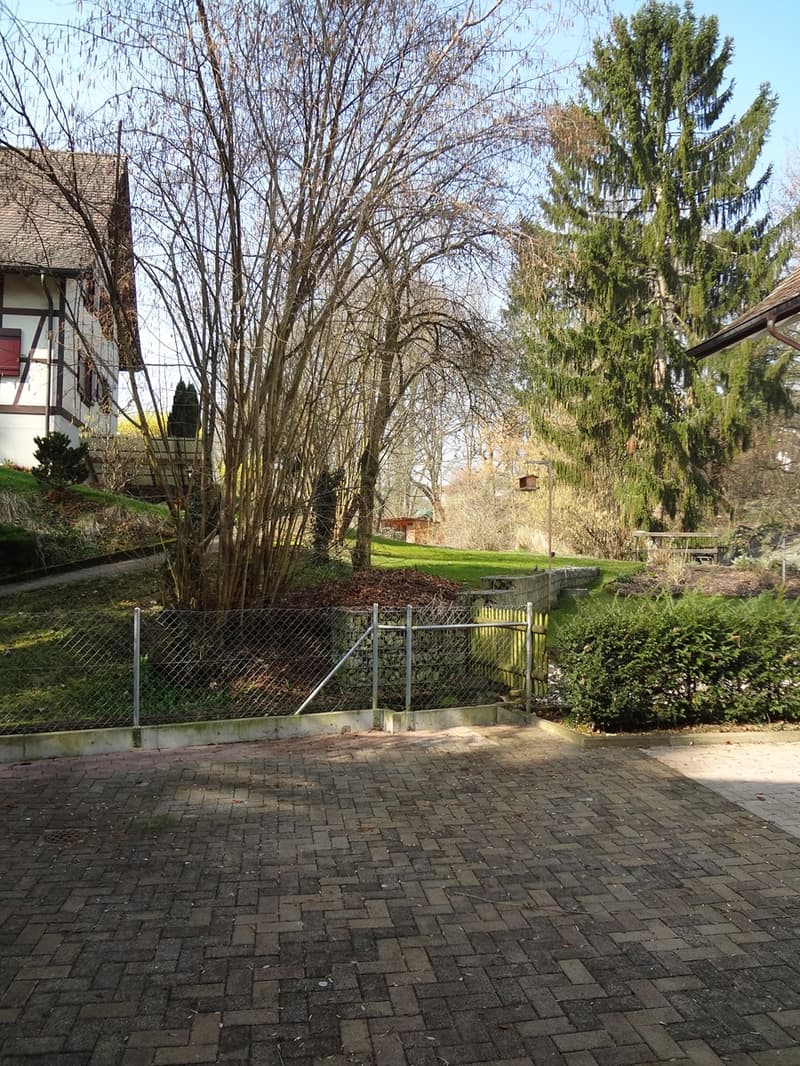 Brick paved ground, fenced entrance, large trees, house in the back, benches in the distance
