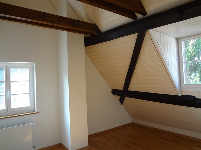 Unfurnished room with wooden beams, slanted ceiling, two windows, and radiator on wall.