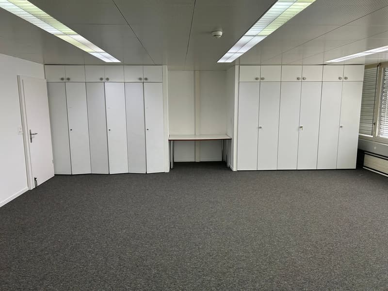 The image shows a large open office space with gray carpeted flooring and white walls. There are several rows of lockers or storage cabinets lining the walls, and a small desk or counter in the center of the room. The ceiling is equipped with fluorescent