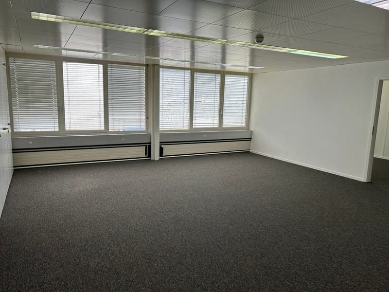 The image shows a large, open office space with gray carpeted flooring, white walls, and large windows with blinds. The ceiling has recessed lighting fixtures. The room appears to be empty, ready for use as an office or workspace.