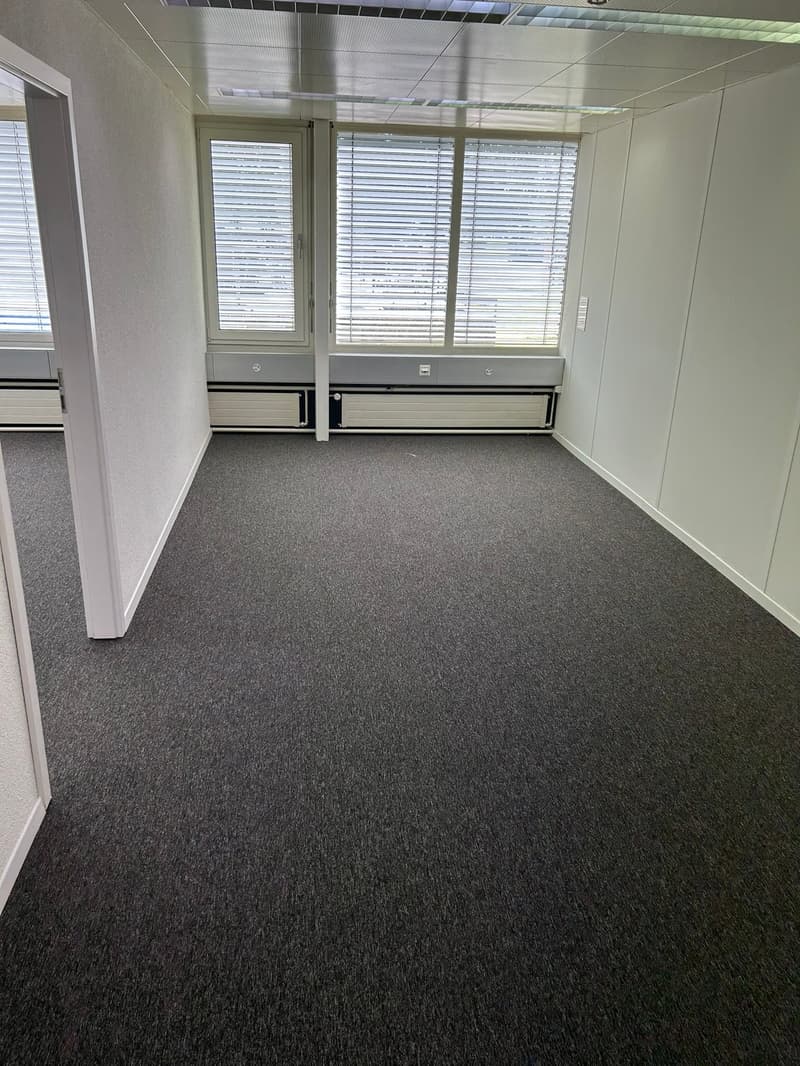 The image shows an empty office space with gray carpeted flooring, white walls, and large windows with blinds. The room appears to be a typical office environment, ready to be furnished and occupied.