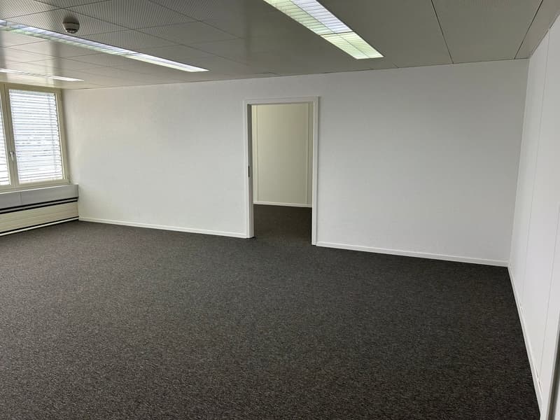 The image shows a spacious office space with white walls, a carpeted floor, and recessed lighting. The room has a large window and a doorway leading to another room. The space appears to be clean, well-lit, and ready for use as an office or workspace.