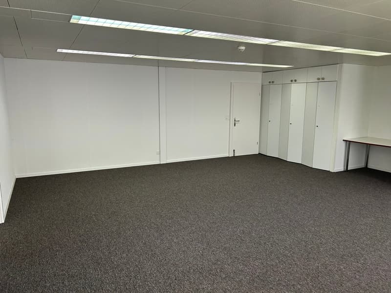 The image shows a spacious office space with gray carpeted flooring, white walls, and recessed lighting fixtures in the ceiling. There are three white doors along the wall, likely leading to other rooms or offices. The room appears to be empty, with no fu
