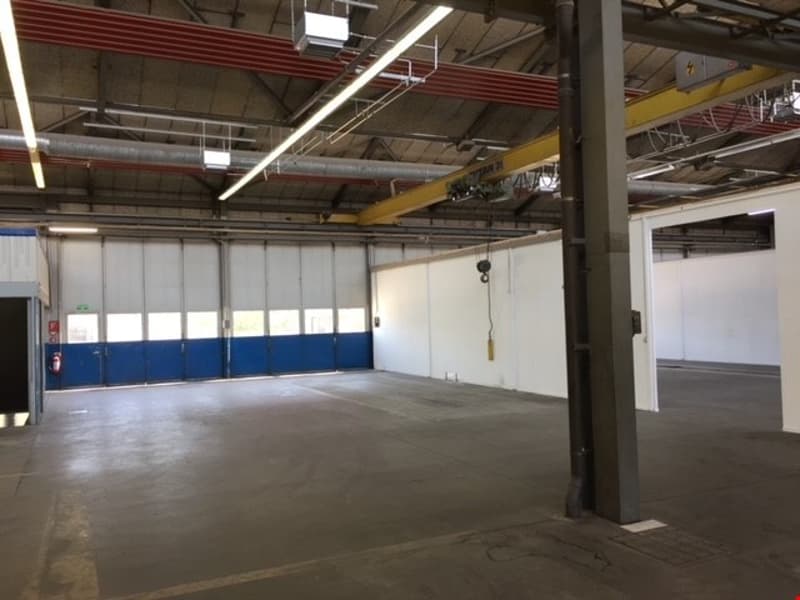 Large open space with concrete floor, metal beams and supports, and overhead lighting. The space appears to be a warehouse or industrial facility with garage doors visible.