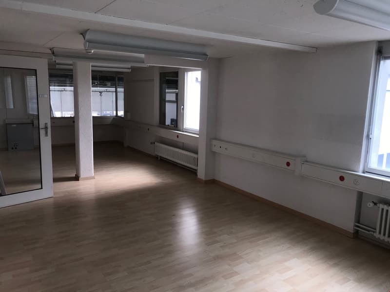 The image shows a large, open office space with a concrete floor, white walls, and overhead lighting fixtures. The room appears to be empty, with no furniture or other equipment visible. There are several windows along one wall, providing natural light. T