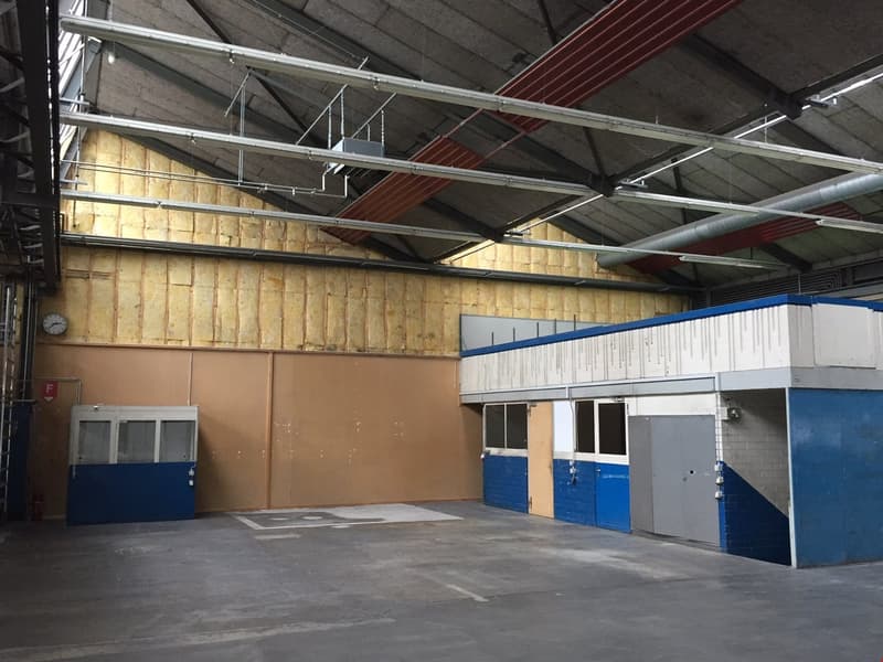 Large open warehouse space with concrete floor, exposed ceiling beams and insulation, blue and white walls, and storage units or offices along the side walls.