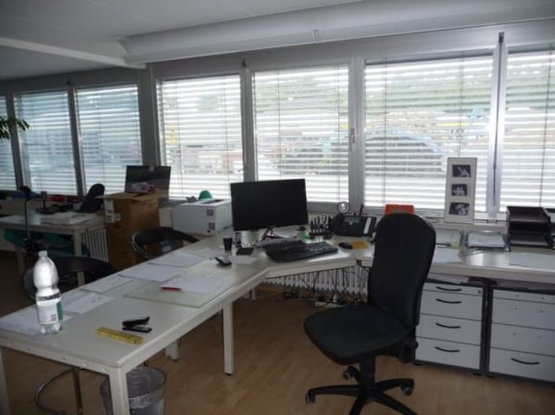 Spacious office with large windows providing a view, multiple desks and workstations, office chairs, and various office equipment and supplies.