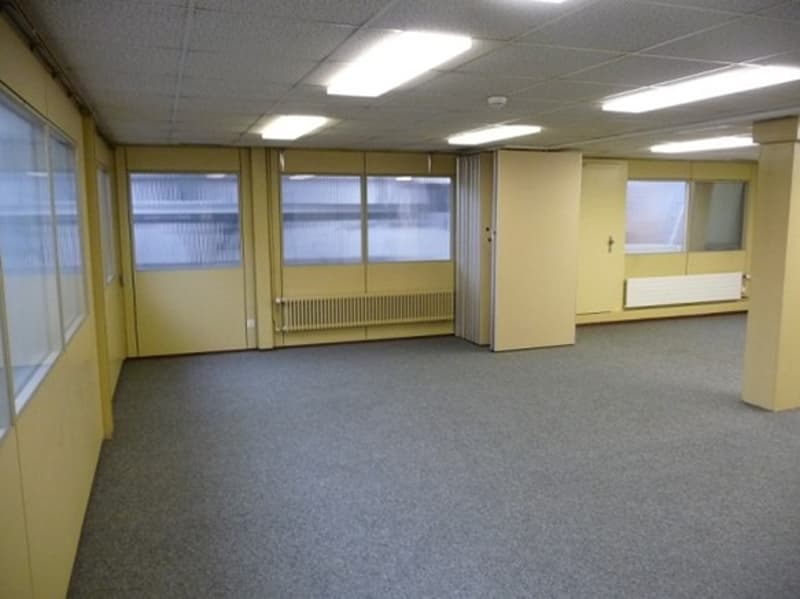The image shows a large, open office space with yellow walls, a tiled floor, and several windows. The room appears to be empty, with no furniture or equipment visible. The space seems to be designed for office use, with a clean and professional appearance