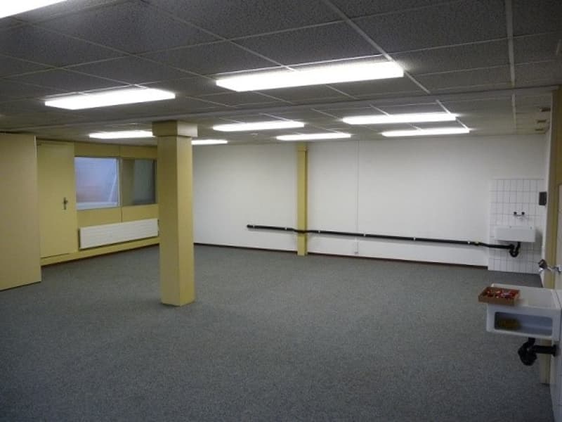 Large open room with gray carpeted floor, white walls, and recessed lighting. The room appears to be an office or commercial space, with a window visible in the background.