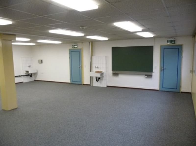 The image shows a classroom-like setting with a chalkboard, desks, and a water fountain. The room has a carpeted floor and fluorescent lighting overhead.