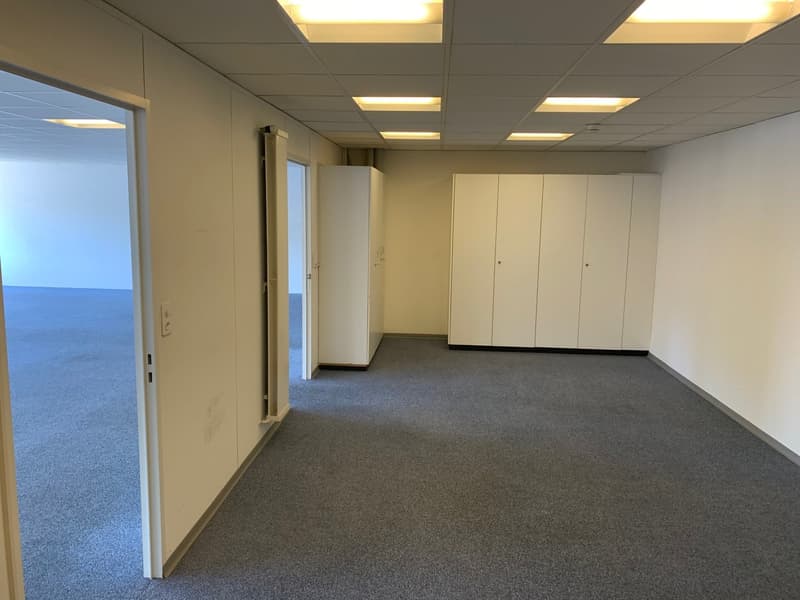 The image shows a spacious office space with a carpeted floor, white walls, and recessed lighting. The room has a large window overlooking an outdoor area. There are several white storage cabinets or lockers along one wall, and the space appears to be set