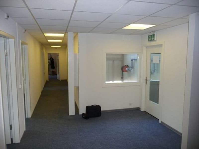 The image shows a hallway in an office space. The hallway has white walls, a blue carpeted floor, and recessed lighting in the ceiling. There are several doors leading to other rooms or offices along the hallway.
