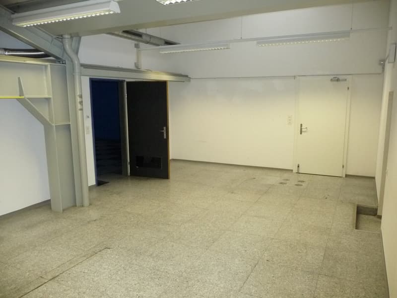 Large open space with concrete flooring, white walls, and overhead lighting. There are shelves and storage units along the walls, and a door leading to another room.