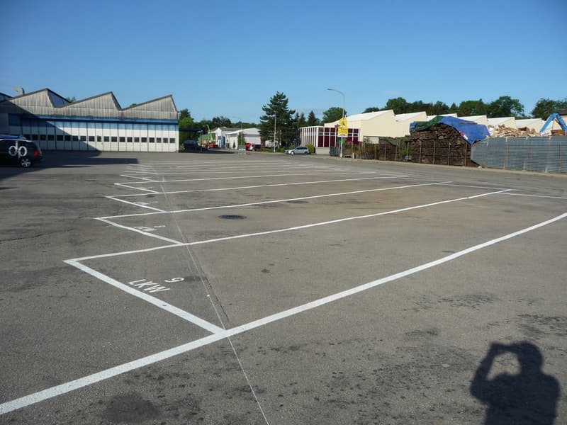 Large paved parking lot with white painted lines marking the parking spaces. Several commercial buildings and structures visible in the background.