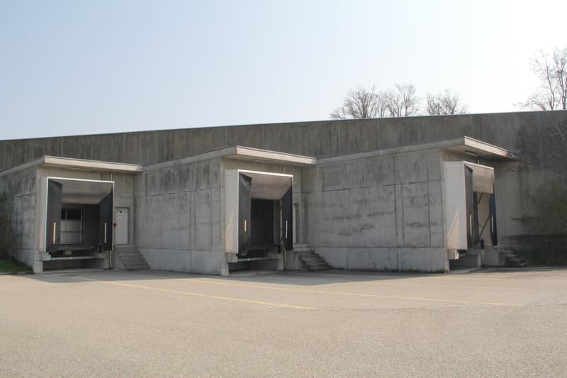 Large concrete warehouse with three garage doors, concrete walls and stairs leading to doors, open parking lot in front