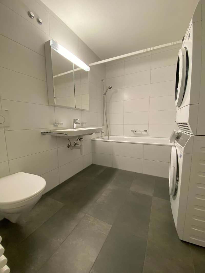 white tiled walls, white tiled floor, bathtub, white toilet, washbasin, mounted mirror, shower, washing machine and dryer