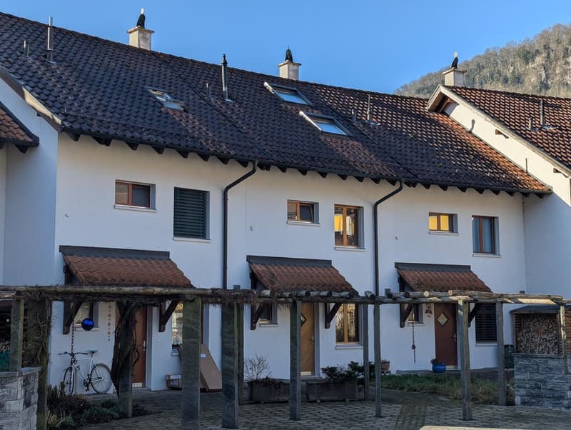 Row of white houses with brown roofs, each with a chimney, brown awnings, and windows with shutters. The houses are surrounded by a brick walkway and have a bicycle parked in front.