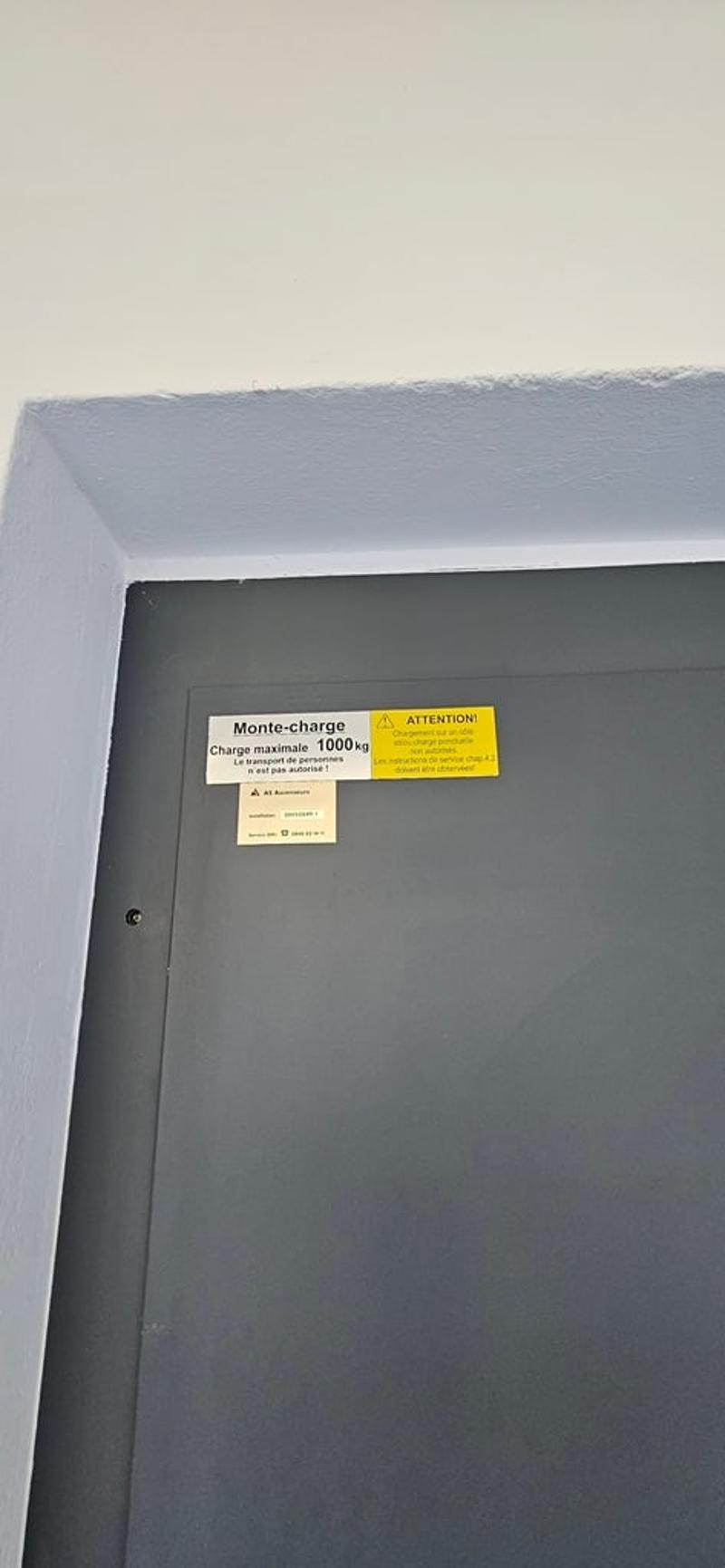 A gray door with two stickers, one yellow and one white, attached to it. The yellow sticker reads 'Attention' and 'Charge maximale 1000kg'. The white sticker reads 'Monte-charge' and 'La valise doit être placée à l'intérieur'.