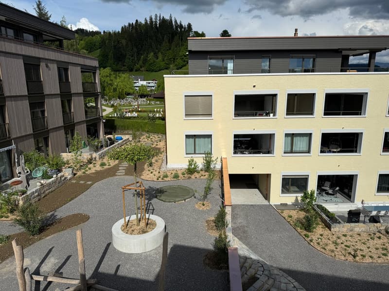 Multi-story residential building with a mix of wooden and yellow exterior, surrounded by landscaped gardens and pathways, with balconies visible on some units.