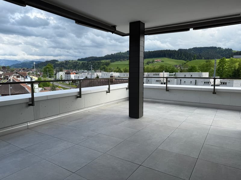 Large balcony with concrete floor tiles, black metal railings, and a view of the surrounding city and mountains.