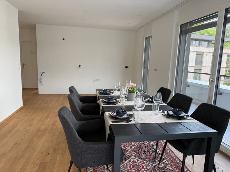 The image shows a modern and minimalist dining room with a large wooden dining table and six black upholstered chairs. The room has large windows that provide natural light and a view of the outdoor greenery. The table is set with plates, glasses, and a f