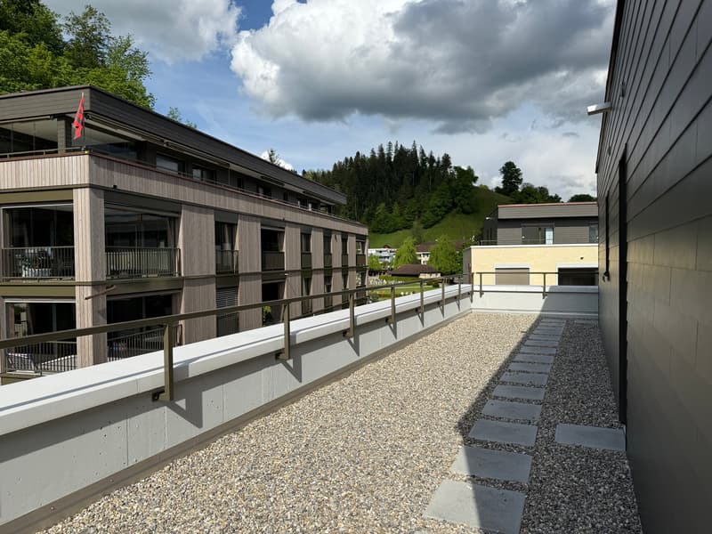 Multi-story building with wooden siding, balconies, and a gravel walkway leading to other buildings. The building is surrounded by trees and a cloudy sky in the background.