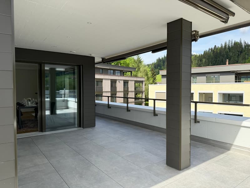 Spacious balcony with glass sliding doors, grey tile flooring, black metal support columns, view of surrounding buildings and trees