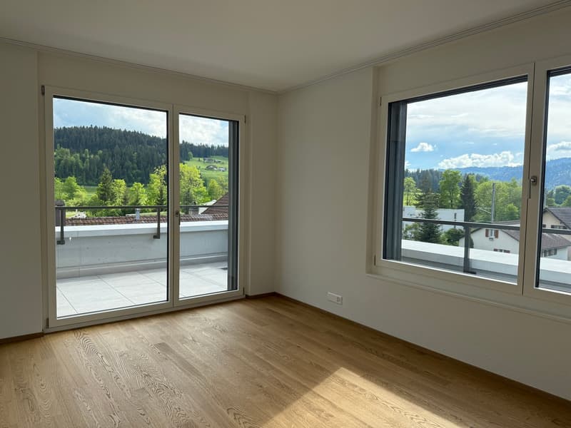 Spacious bedroom with large windows offering a scenic view of the surrounding forest and mountains. The room has hardwood flooring and a sliding glass door leading to a balcony or terrace.
