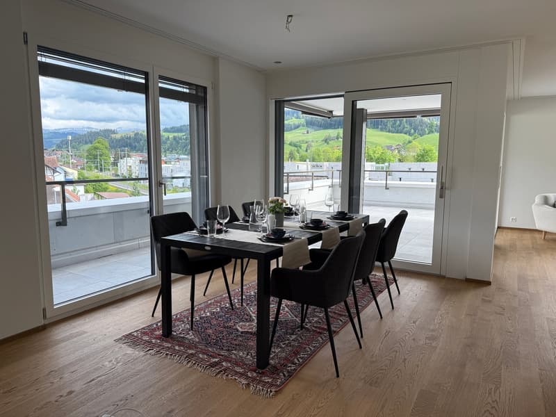 Spacious dining room with large windows offering a scenic view of the surrounding countryside. The room features a modern black dining table with chairs, a decorative rug, and hardwood flooring. The overall design creates a cozy and inviting atmosphere.