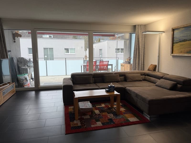 Brown sectional sofa, coffee table, multicolored rug, floor-to-ceiling glass doors, balcony with outdoor furniture, framed picture, side table with lamp