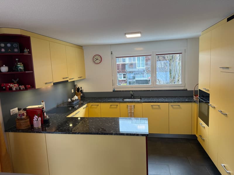 Modern kitchen with yellow cabinets, granite countertops, stainless steel appliances, two windows with blinds, ceiling-mounted light fixture, and various decorative items on the shelves.