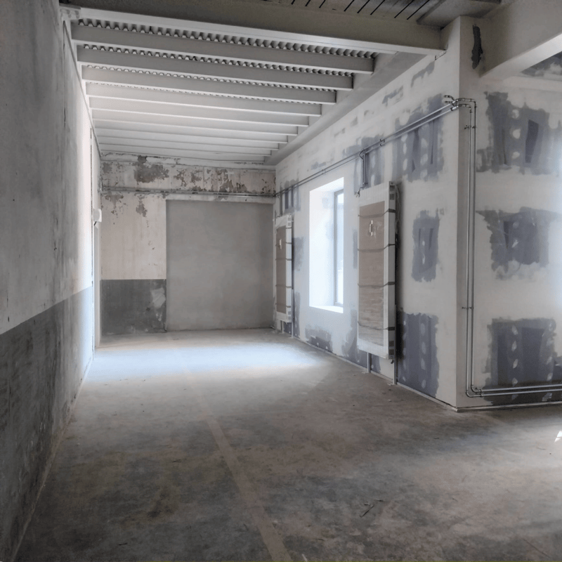This image shows an empty room undergoing renovation. The walls are partially plastered and the floor is bare concrete. The ceiling has exposed beams and supports. The room appears to be in a state of construction or remodeling, with materials and tools v