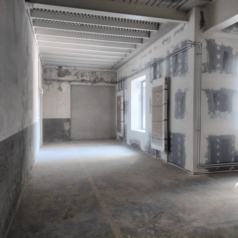 This image shows an empty room undergoing renovation. The walls are partially plastered and the floor is bare concrete. The ceiling has exposed beams and supports. The room appears to be in a state of construction or remodeling, with materials and tools v