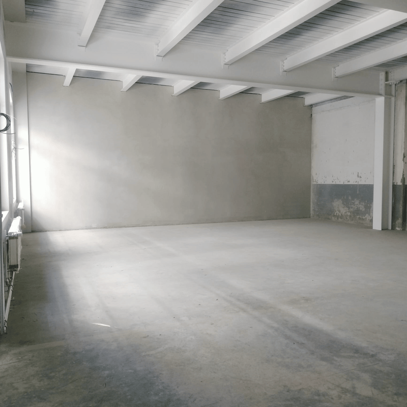 Large empty room with white walls and a high ceiling with exposed wooden beams. The floor appears to be a smooth concrete or cement surface. The room has a spacious and minimalist feel, suggesting it could be used as a studio, workshop, or storage space.