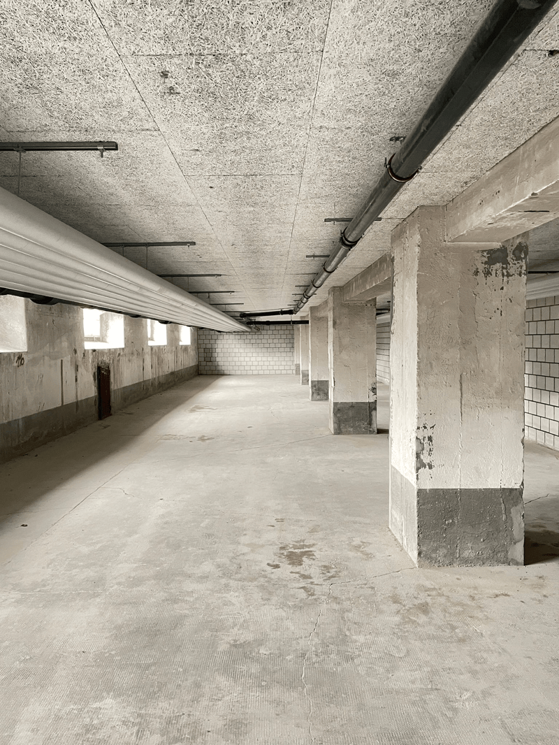 Wide, empty hallway, concrete floor, pipes on the ceiling, white walls, columns, and brick wall.